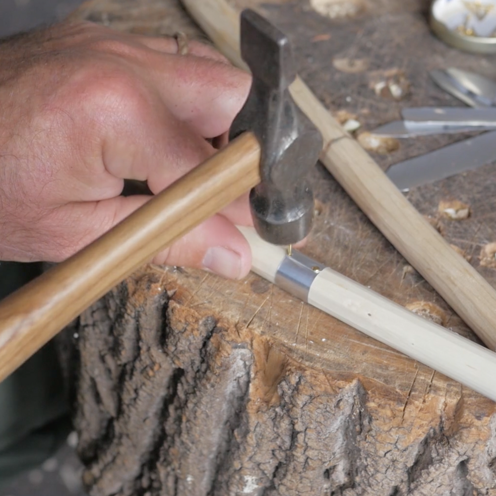 A person is shaping a beige hazel stick with a hammer on a wooden anvil that is attached to a larger piece of wood, likely a workshop demonstration or practice.