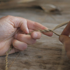 A close up of hands making string using tī kōuka (cabbage tree) leaf resting on a brown wooden table.