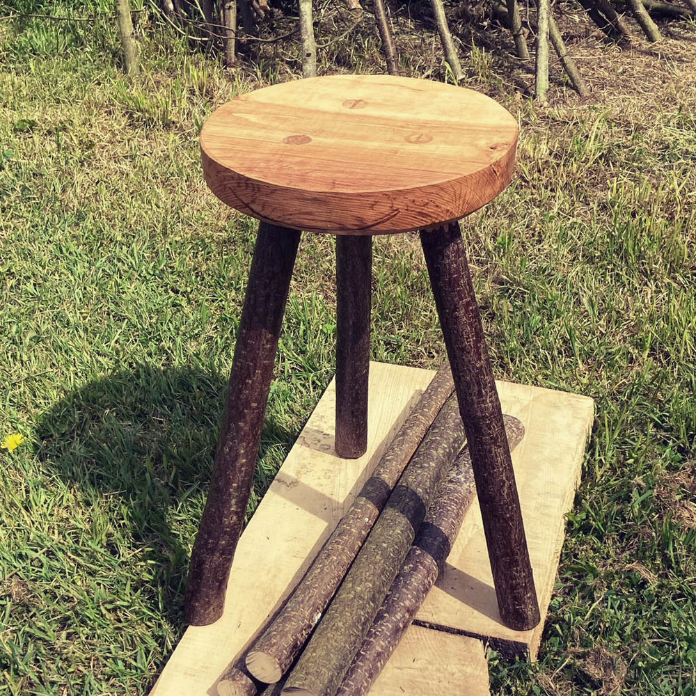 Three legged wooden stool on a wooden platform on some grass outside.