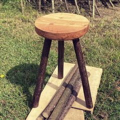 Three legged wooden stool on a wooden platform on some grass outside.