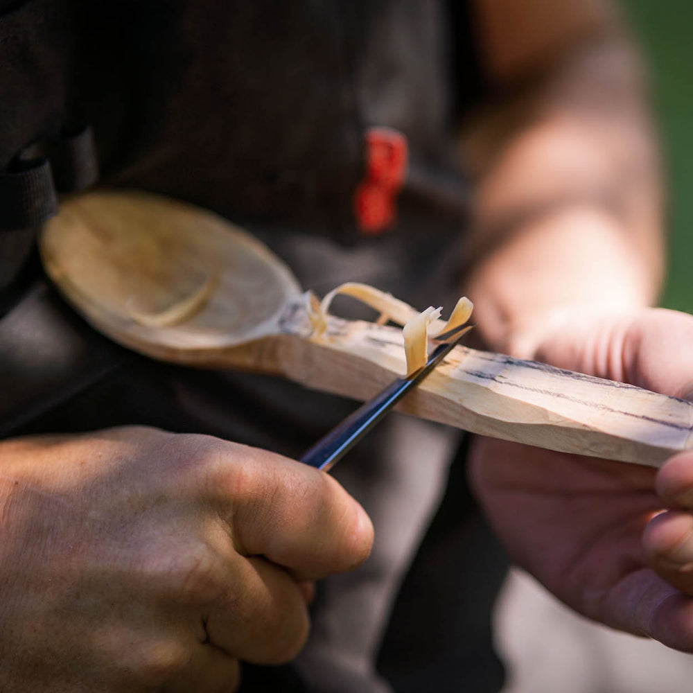 Person using a sharp knife to carve a wooden spoon outdoors