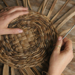 A person's hands weaving a round placemat made of brown tī kōuka leaves on a wooden surface with a tool.