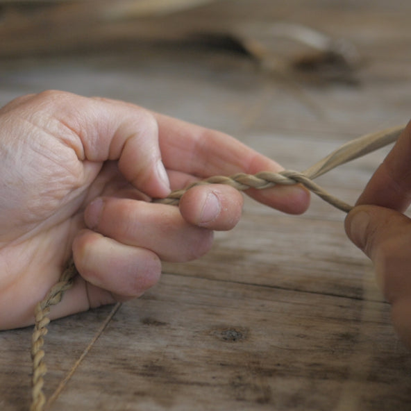 A close up of hands making string using tī kōuka (cabbage tree) leaf resting on a brown wooden table.